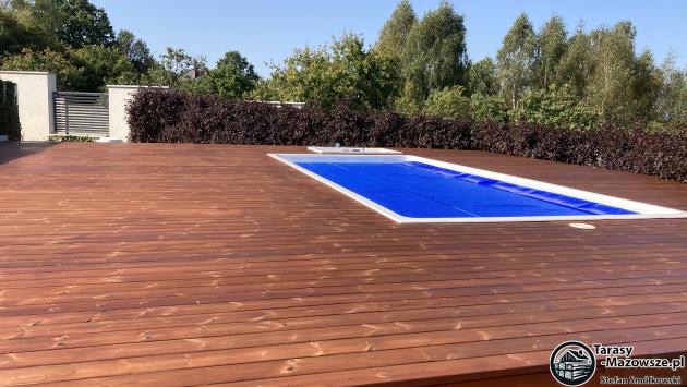 Rozległy taras z desek Thermory Thermo sosna, wykończony olejem do tarasów, otaczający basen przykryty niebieską plandeką, z widoczną konstrukcją krzyżową i fundamentami punktowymi, w tle zielona...