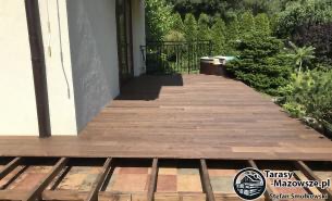 Brązowy taras drewniany na konstrukcji z częściowo odsłoniętą kostką brukową, widok z góry, dom z jasną elewacją w tle.