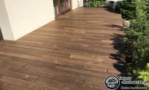 Brązowy taras z desek, widok z góry, z elementami zieleni w tle oraz z widocznym fragmentem budynku i balustrady.