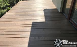 Nowoczesny taras z desek w odcieniach brązu, ułożonych poziomo, z widocznym cieniem padającym na część powierzchni, przylegający do budynku z jasną elewacją i ciemnymi drzwiami.