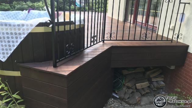 Fragment tarasu z desek kompozytowych w odcieniach brązu, z metalową balustradą o ozdobnych elementach, przylegający do budynku z widocznym składem drewna pod spodem.