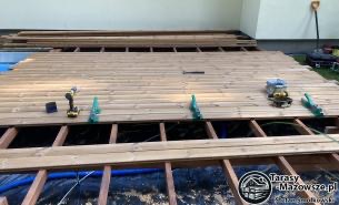 Montaż drewnianego tarasu: widok na konstrukcję z legarów i częściowo ułożone deski, w tle widoczny budynek i zieleń, na pierwszym planie wkrętarka i poziomica.