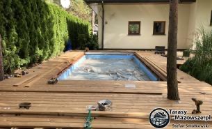 Budowa drewnianego tarasu wokół basenu w Warszawie, widoczne deski w trakcie montażu i narzędzia pracy. W tle zielony żywopłot.
