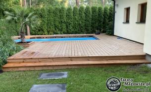 Jasny taras z drewna egzotycznego otaczający basen, z palmą w donicy, widok z poziomu trawnika, w tle żywopłot z tui.