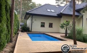 Nowoczesny taras z jasnego drewna otaczający basen na tle domu z ciemną dachówką i zielenią drzew.
