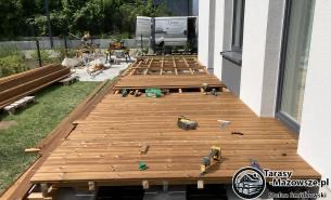 Montaż desek na drewnianej konstrukcji tarasu przylegającego do budynku. Widoczne narzędzia, poziomica i wkrętarka. W tle trawa i ogrodzenie.