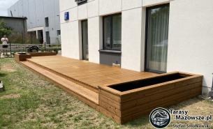 Drewniany taras z dwoma stopniami, zintegrowany z donicami po bokach, na tle białej elewacji budynku. Widoczne nierówności trawnika.