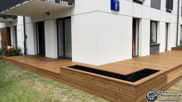 Nowoczesny taras z desek w odcieniach brązu, przylegający do białego budynku z czarnymi ramami okiennymi i numerem 8 na niebieskiej tabliczce, z wbudowaną drewnianą donicą wyłożoną czarną folią.