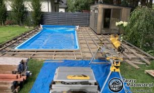 Budowa tarasu przy basenie: widoczne legary na trawie, basen przykryty plandeką, w tle domek narzędziowy i ogrodzenie. Na pierwszym planie piła stołowa i ukośnica na statywie.