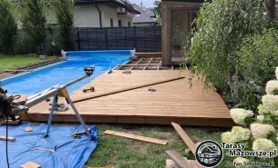 Budowa drewnianego tarasu przy basenie, widoczne deski, konstrukcja nośna, narzędzia i osłonięty basen w tle.