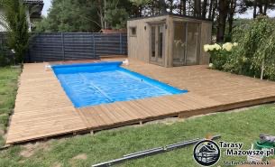 Jasny, drewniany taras okalający basen z niebieską wodą, w tle mała drewniana altana z przeszklonymi drzwiami i oknem, całość otoczona zielenią trawnika i drzew.