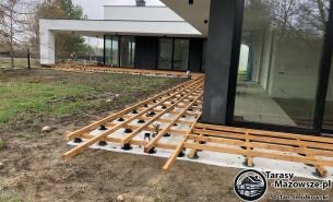 Konstrukcja pod przyszły taras przy nowoczesnym domu z dużymi oknami. Widoczne legary na regulowanych wspornikach, przygotowane do montażu desek tarasowych. W tle trawnik i drzewa.