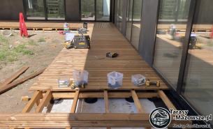 Montaż drewnianego tarasu: widoczne legary, deski w trakcie układania, narzędzia pracy (wiertarka, poziomica), częściowo ułożona powierzchnia, widok z boku budynku z dużymi oknami.