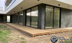 Nowoczesny taras z jasnego drewna przy budynku z dużymi oknami, widoczne elementy konstrukcyjne pod tarasem, w tle trawnik i budynek, logo firmy w prawym dolnym rogu.