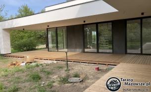 Nowoczesny taras drewniany z jasnego drewna przy białym domu z dużymi oknami przesuwnymi, widoczne niedokończone prace i materiały budowlane na trawniku.