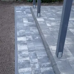 Świeżo ułożona szara kostka brukowa z obrzeżem, widoczne słupy drewnianej konstrukcji, ziemia przygotowana pod nasadzenia, w tle zieleń i żółte kwiaty.