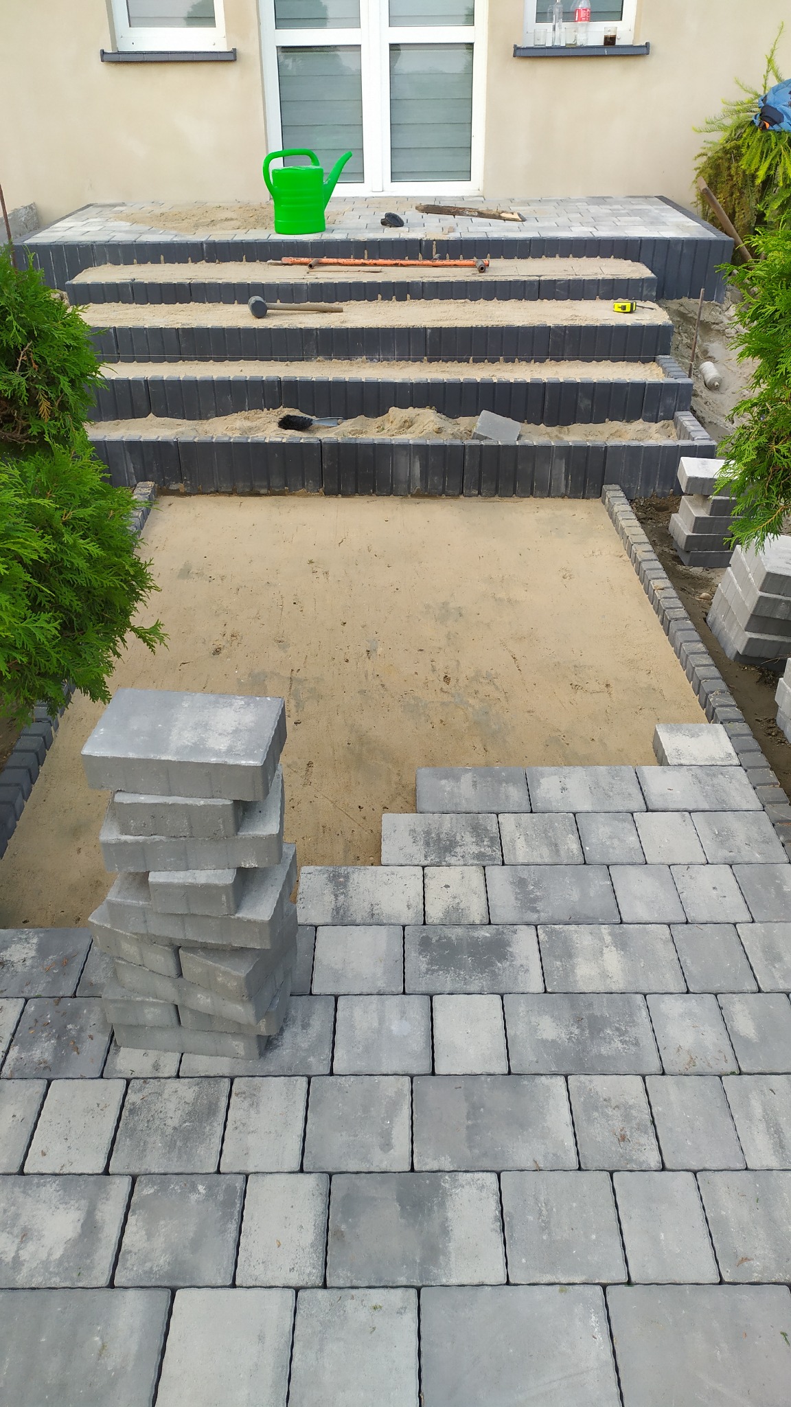 Montaż stopni i nawierzchni z szarej kostki brukowej przed wejściem do domu, widoczna zielona konewka i przygotowane podłoże z piasku.