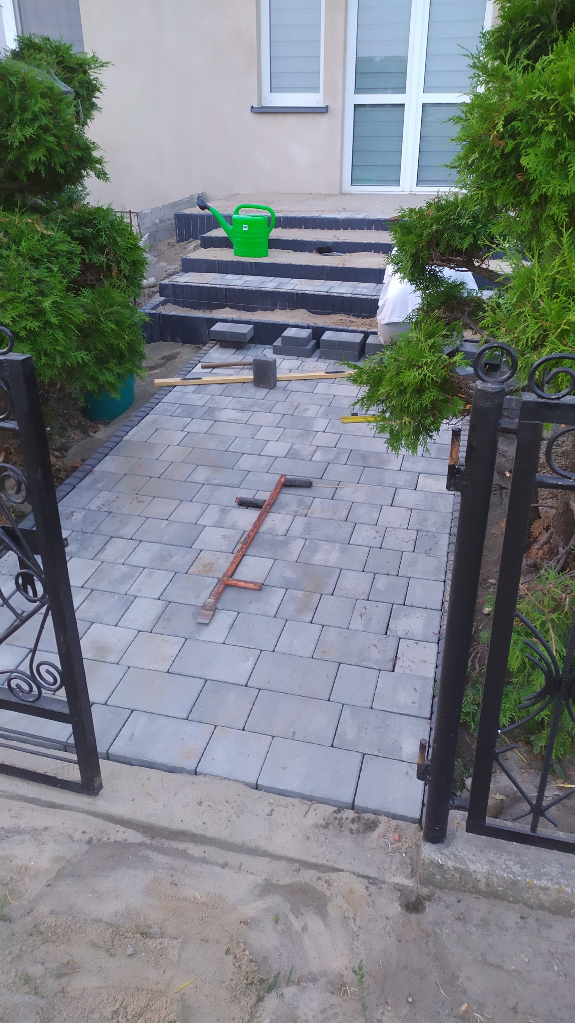 Szara kostka brukowa ułożona na podjeździe, widoczny fragment metalowej bramy, zielona konewka na schodach z kostki, narzędzie do układania kostki leżące na bruku.