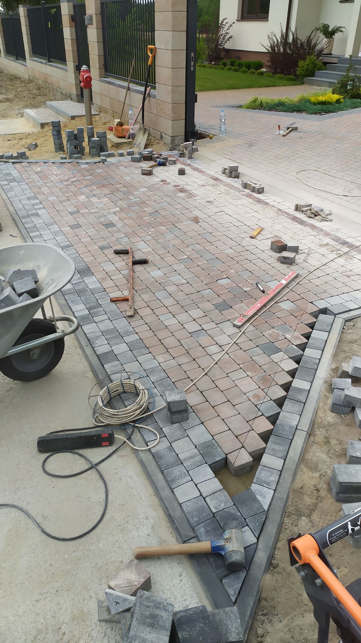 Fragment podjazdu w trakcie prac brukarskich, widoczne ułożone kostki w odcieniach szarości i brązu, narzędzia brukarskie, taczka z kostką oraz przygotowane podłoże pod dalszą część nawierzchni.