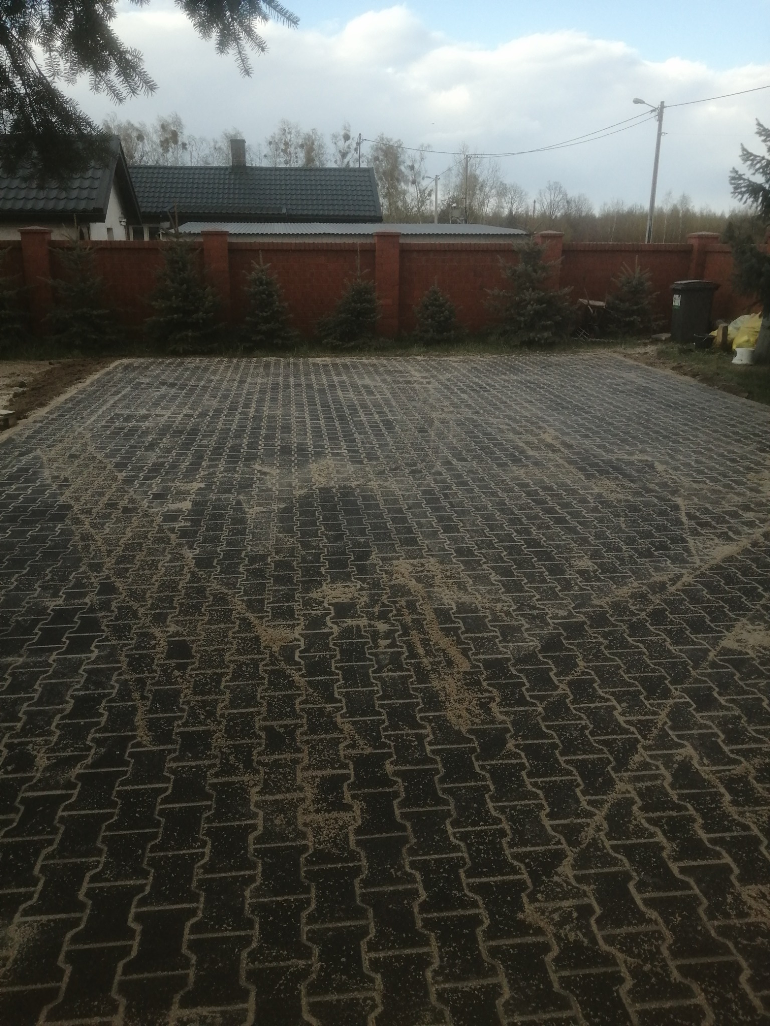 Ciemna kostka brukowa ułożona na posesji, zasypana piaskiem, widoczny fragment domu z ceglanym ogrodzeniem i młodymi drzewkami iglastymi w tle.