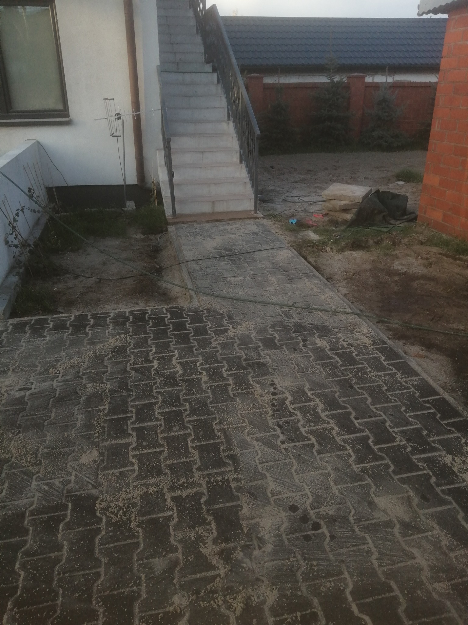 Nowo ułożona, szara kostka brukowa typu 'kośc' prowadząca od schodów wejściowych do domu, posypana piaskiem do wypełnienia szczelin, widok z góry.