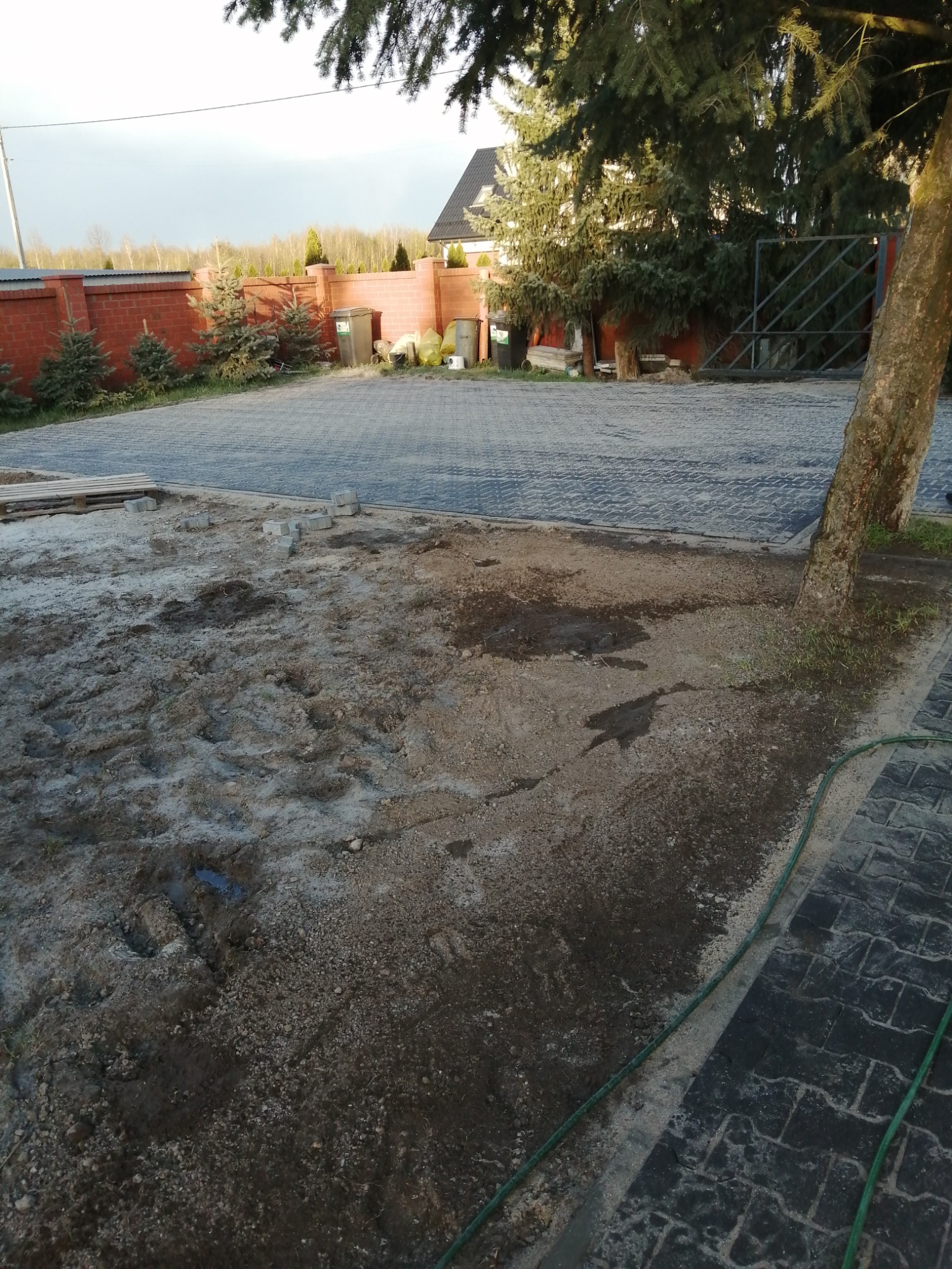 Układanie kostki brukowej: widok na częściowo ułożoną szarą kostkę brukową, z przygotowanym podłożem i stosem kostek do ułożenia, na tle zieleni i ogrodzenia z czerwonej cegły.