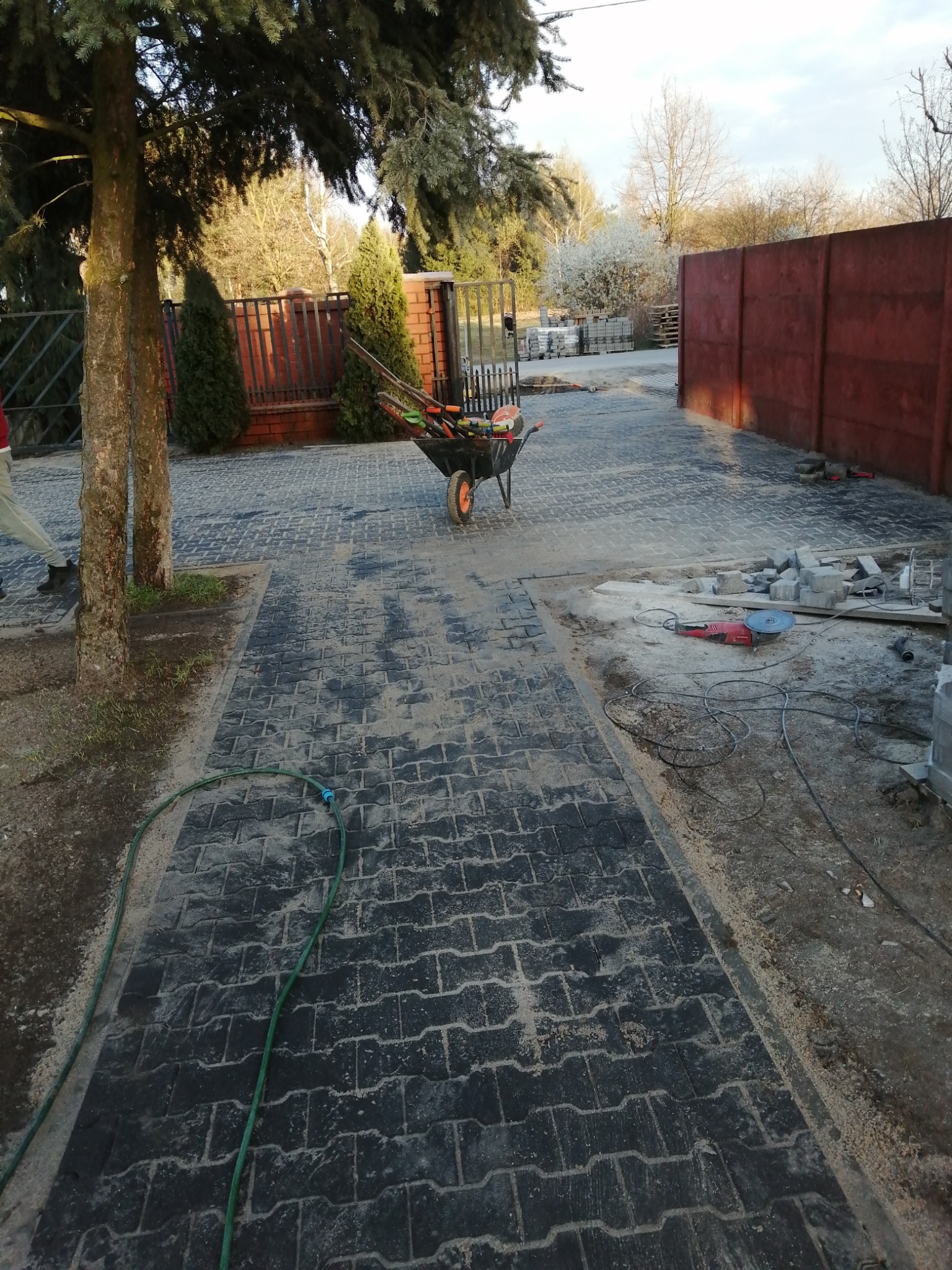 Ciemna kostka brukowa w trakcie układania, widoczna piła do cięcia, wózek z narzędziami, zielony wąż ogrodowy i fragment ogrodzenia w tle.