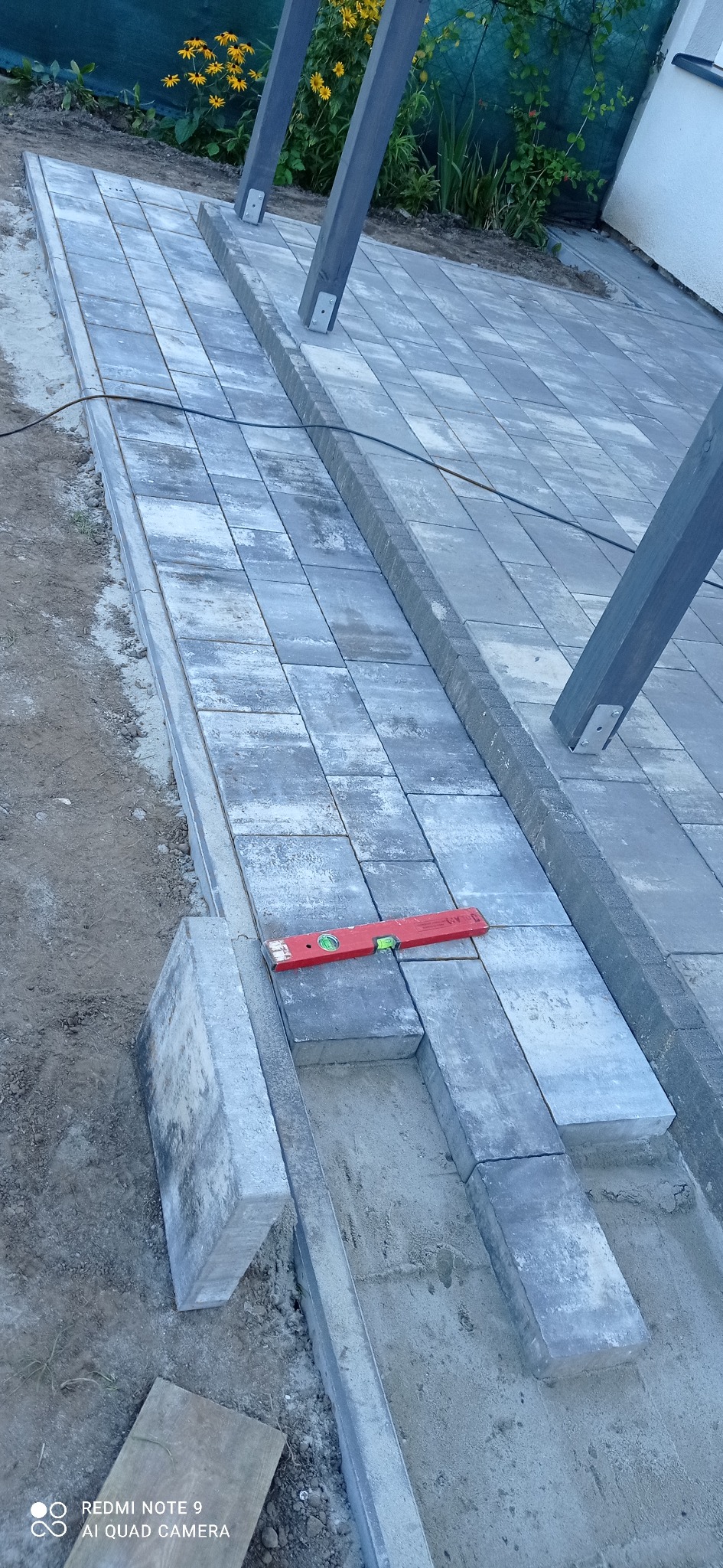 Ułożona szara kostka brukowa z poziomnicą w trakcie prac, widoczne elementy konstrukcji z drewna i fragment ogrodu z kwiatami w tle.