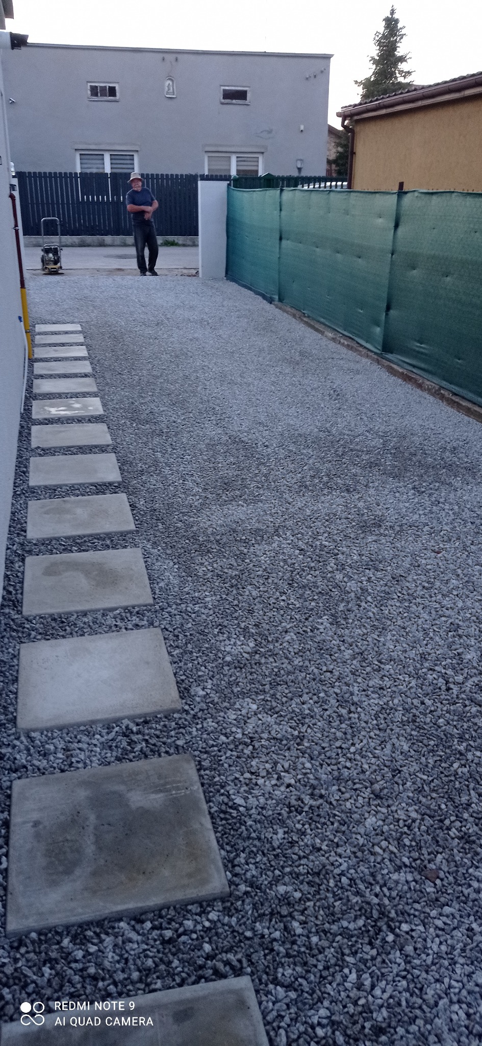 Wykończona ścieżka z betonowych płyt ułożonych w szarym żwirze, z widocznym pracownikiem w tle i ubijarką wibracyjną, oddzielona od sąsiada zieloną siatką.