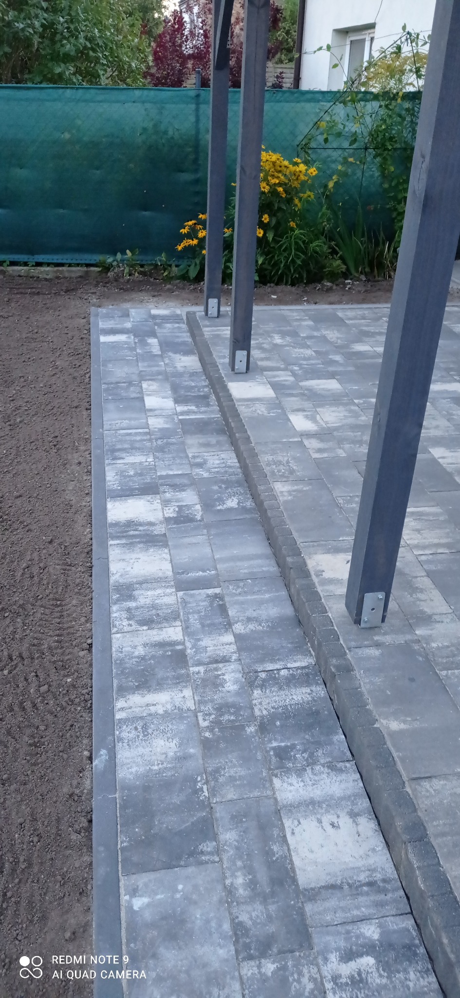 Świeżo ułożona szara kostka brukowa z obrzeżem, widoczne słupy drewnianej konstrukcji, ziemia przygotowana pod nasadzenia, w tle zieleń i żółte kwiaty.