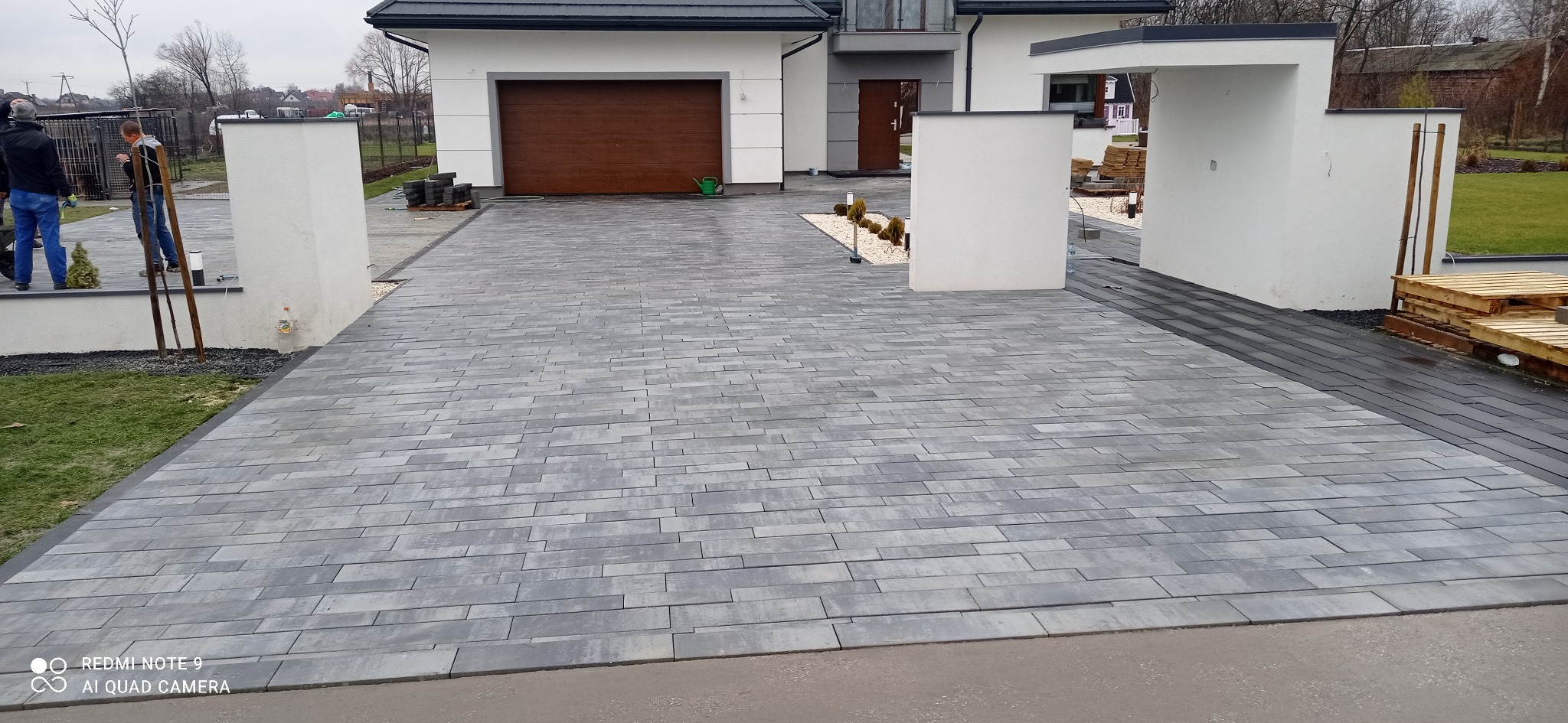 Ułożona szara kostka brukowa w kształcie prostokątów na podjeździe przed nowoczesnym domem z brązową bramą garażową, widoczne białe słupki i elementy dekoracyjne z kamyków.
