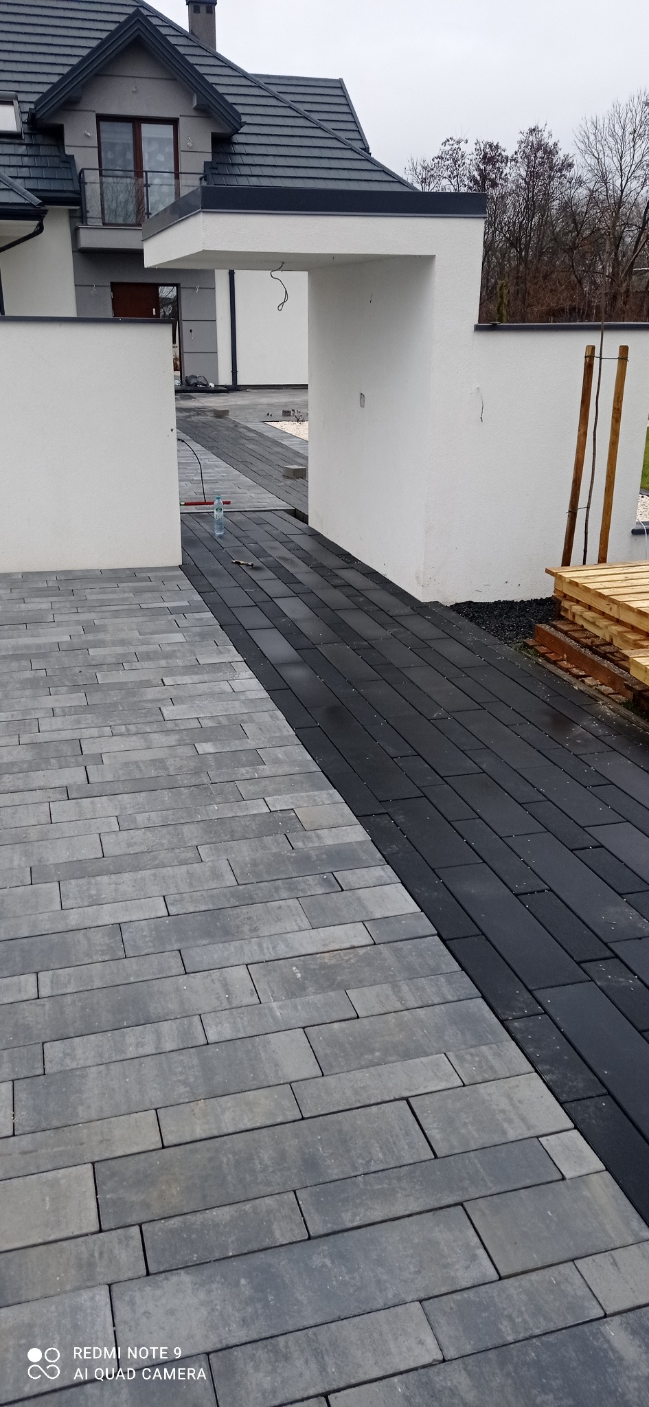 Ułożona kostka brukowa w dwóch kolorach - szarym i czarnym, prowadząca przez białą bramę do domu, widoczna butelka wody i palety na boku.