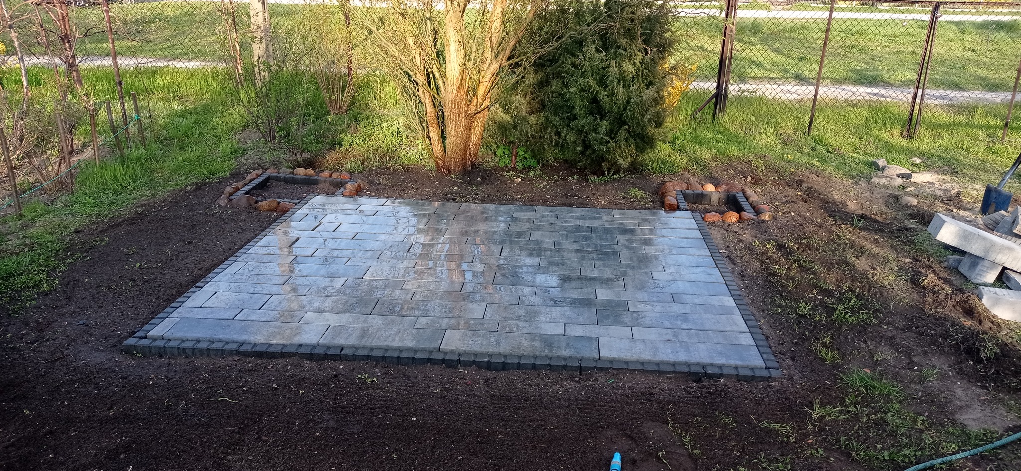 Ułożona, prostokątna nawierzchnia z szarych, podłużnych elementów w ogrodzie, obrzeża z ciemniejszych kostek, widoczne przygotowane podłoże i elementy dekoracyjne z cegieł.
