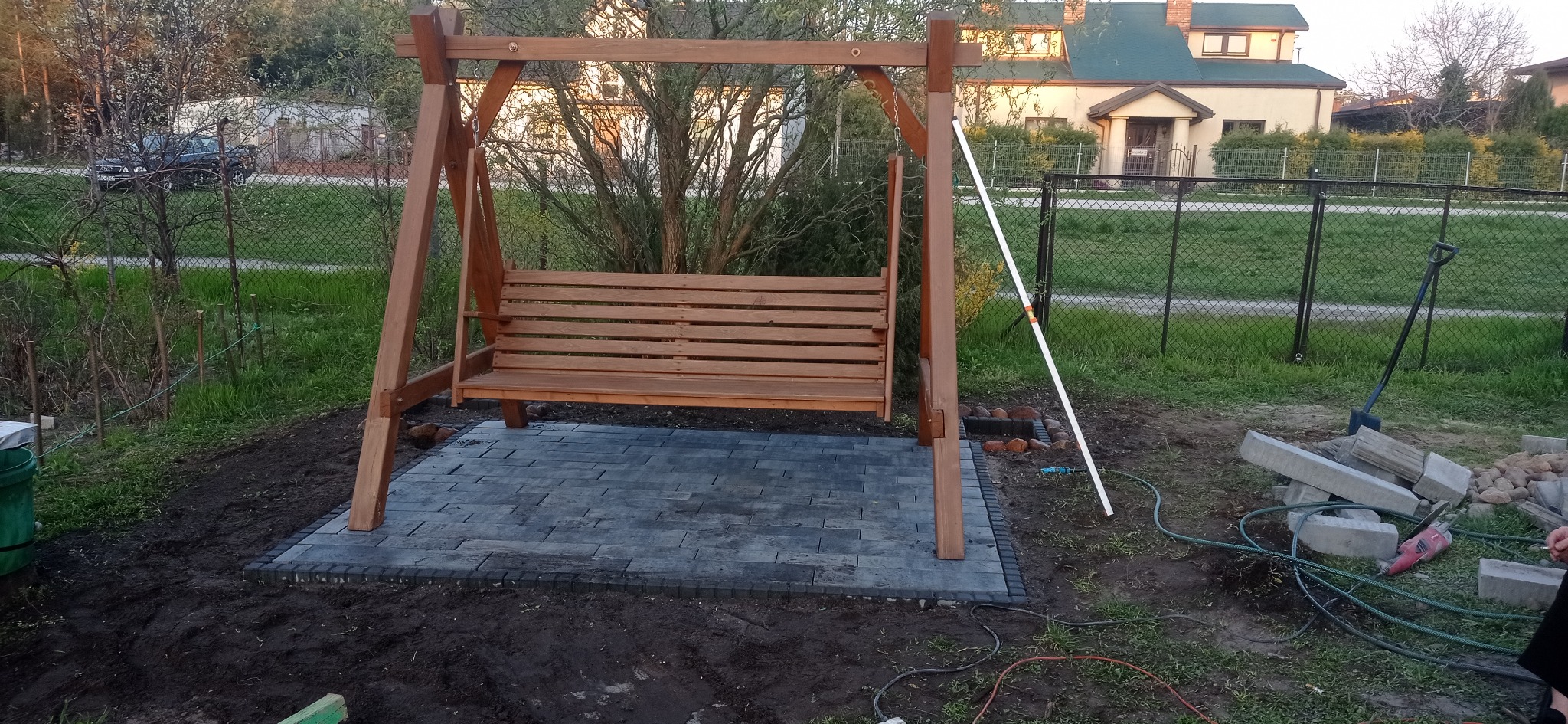 Drewniana huśtawka ogrodowa na szarym brukowanym podłożu, widoczne narzędzia i materiały budowlane, dom w tle.