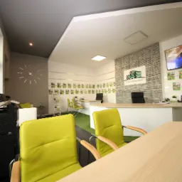 Wnętrze biura agencji nieruchomości z zielonymi akcentami: krzesła i trawa na podłodze, na ścianie logo firmy i zdjęcia ofert, na biurku kalendarz.