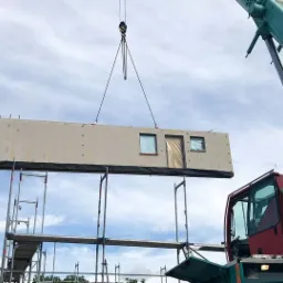 Montaż modułu domu z oknami i drzwiami za pomocą dźwigu na tle nieba z chmurami.