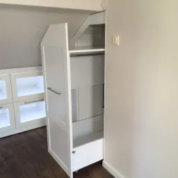 Biała szafa wnękowa z otwartymi drzwiami, widoczna półka, drążek i wysuwana szuflada na kółkach, dopasowana do skosu dachu, obok zabudowa meblowa z przeszklonymi drzwiczkami.