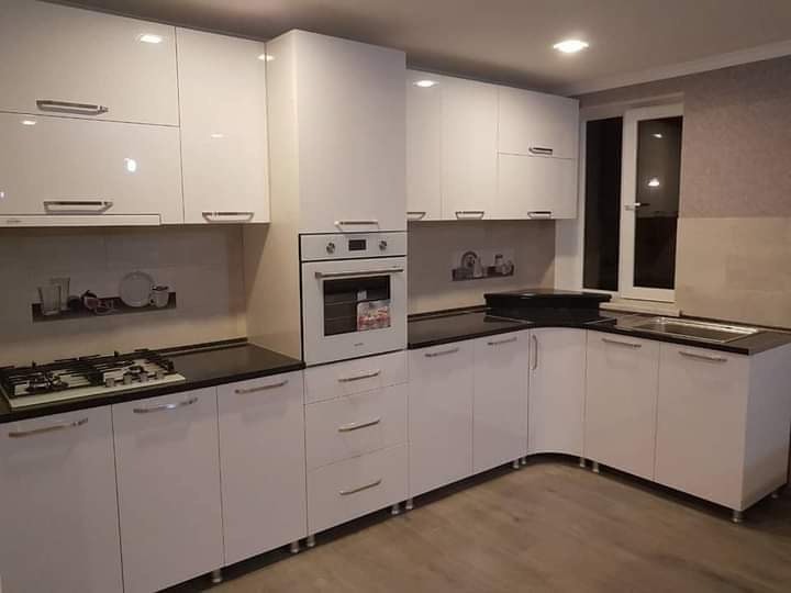 Biała, narożna zabudowa kuchenna z czarnym blatem, widok z niskiego kąta, oświetlenie sufitowe, okno w tle.