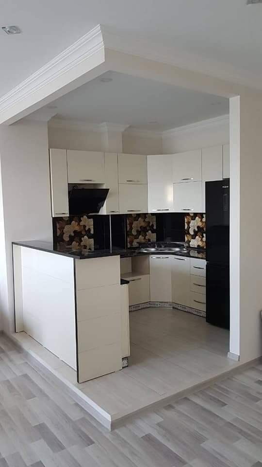 Nowoczesna, biała kuchnia na wymiar z czarnym blatem i panelem ściennym z motywem kwiatowym, zlokalizowana w pomieszczeniu z podwyższoną podłogą i czarną lodówką.