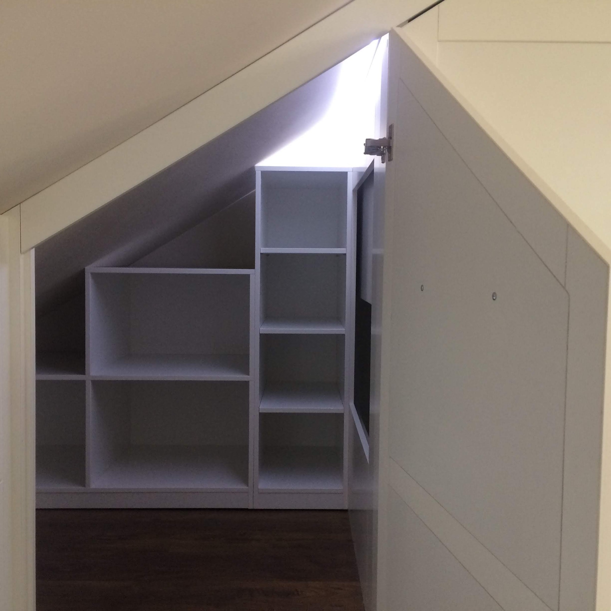 Biała szafa wnękowa z otwartą częścią regałową, dopasowana do skosu dachu, widoczna na tle ciemnej podłogi.