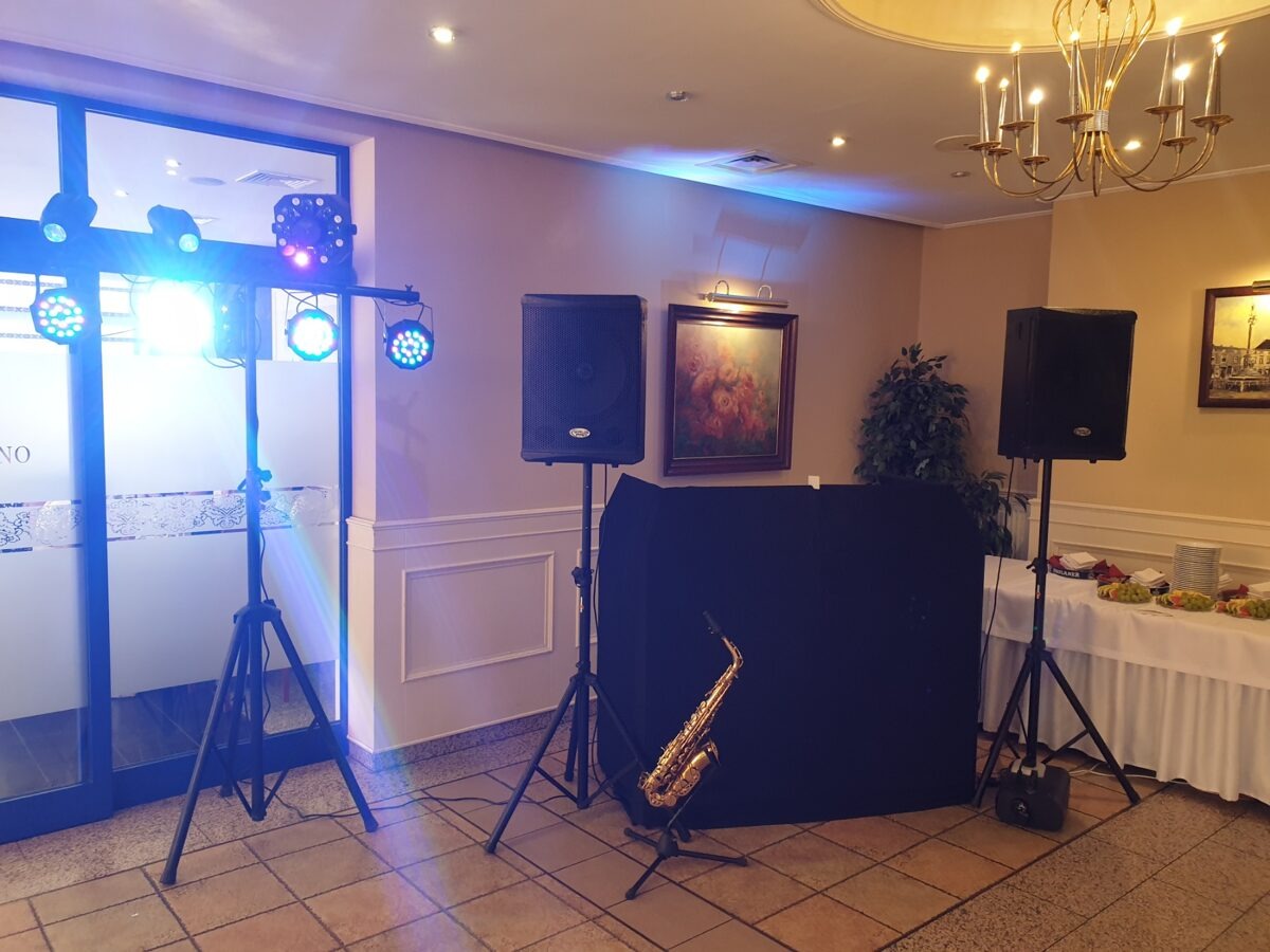 Sala bankietowa przygotowana na imprezę: oświetlenie sceniczne LED na statywie, kolumny głośnikowe na statywach, saksofon na statywie przed czarnym parawanem, stół z przekąskami w tle, żyrandol...