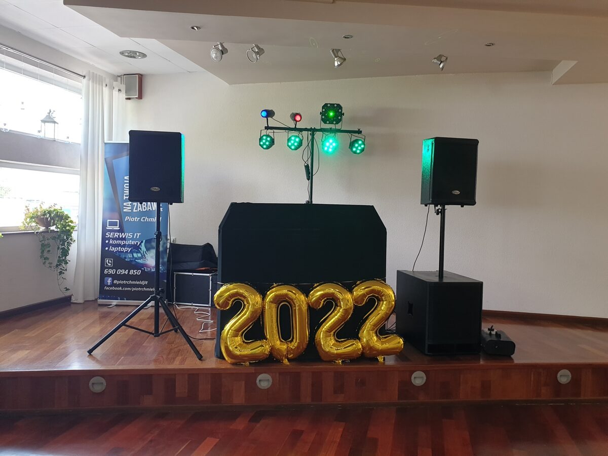 Scena z nagłośnieniem, oświetleniem i konsoletą DJ-ską, udekorowana złotymi balonami w kształcie cyfr '2022', z widocznym banerem reklamowym firmy IT w tle.