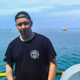 Mężczyzna w czarnej koszulce z logo firmy z Gdańska na tle morza i statków, ubrany w robocze spodnie z kieszeniami.