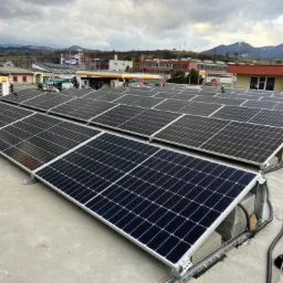 Rząd paneli słonecznych na płaskim dachu budynku z widokiem na zabudowania miejskie i góry w tle pod pochmurnym niebem.