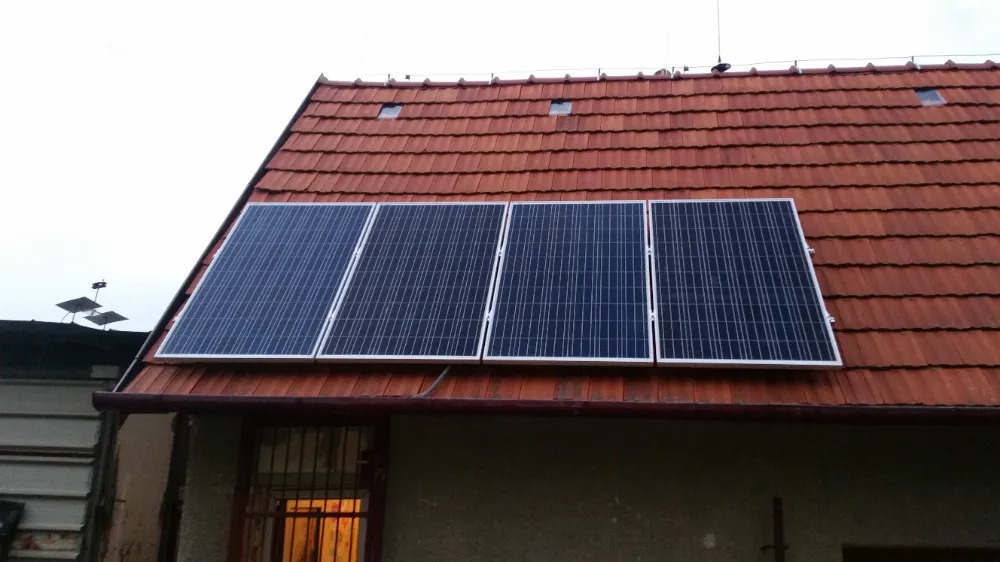 Cztery panele słoneczne zamontowane na czerwonym, dachówkowym dachu budynku z małym oknem dachowym i anteną na szczycie.