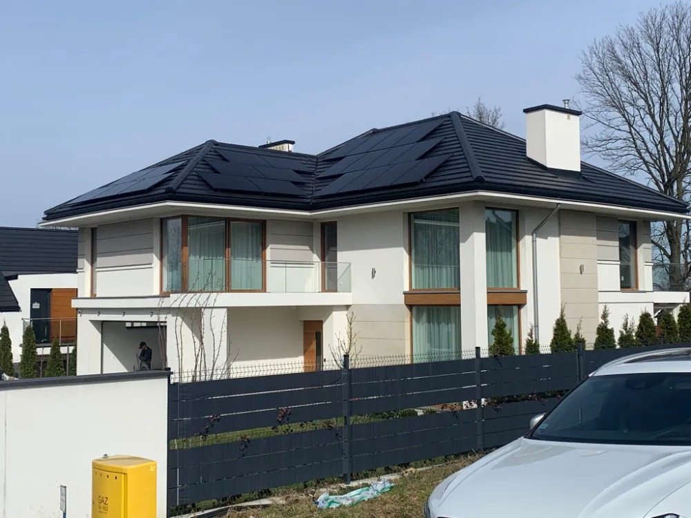 Nowoczesny dom jednorodzinny z panelami słonecznymi na dachu, widok z poziomu ulicy, z przodu zaparkowany biały samochód, w tle drzewa bez liści.