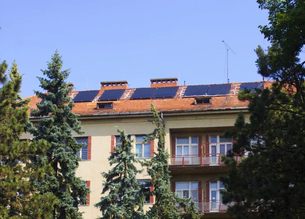 Panele słoneczne zamontowane na dachu budynku z czerwonej dachówki, widoczne zza drzew iglastych, w tle błękitne niebo.