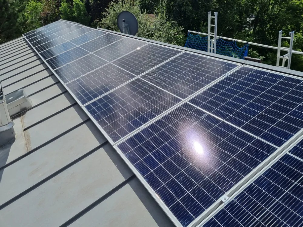 Instalacja paneli słonecznych na metalowym dachu, widoczna drabina zabezpieczona siatką oraz antena satelitarna w tle.