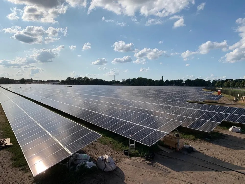 Rozległa farma fotowoltaiczna z panelami odbijającymi światło słoneczne, widoczne drabina i materiały montażowe, błękitne niebo z chmurami w tle.
