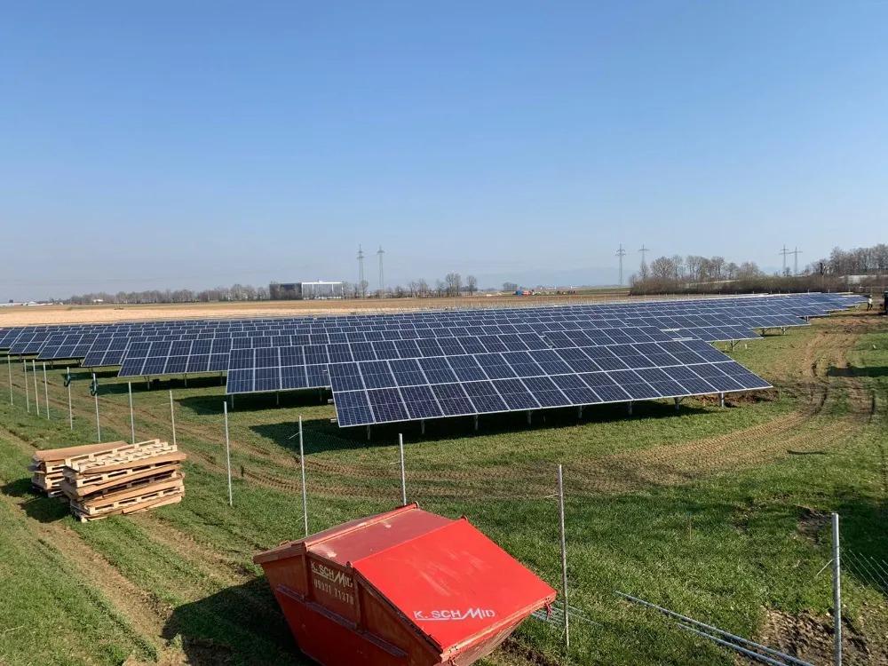 Rozległa farma paneli słonecznych na polu, z czerwonym kontenerem i stertą palet w pierwszym planie, widoczne linie wysokiego napięcia w oddali.